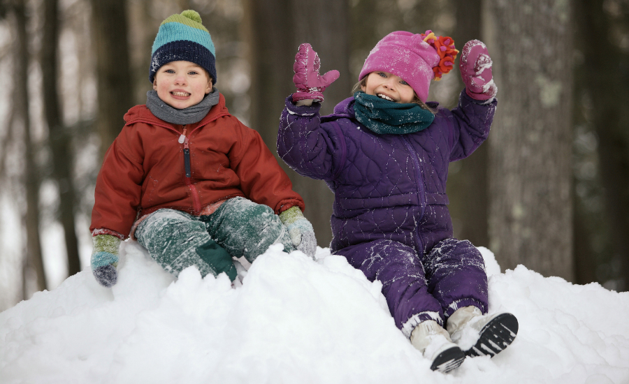 Two children in winter clothes sitting on top of a pile of snow smiling